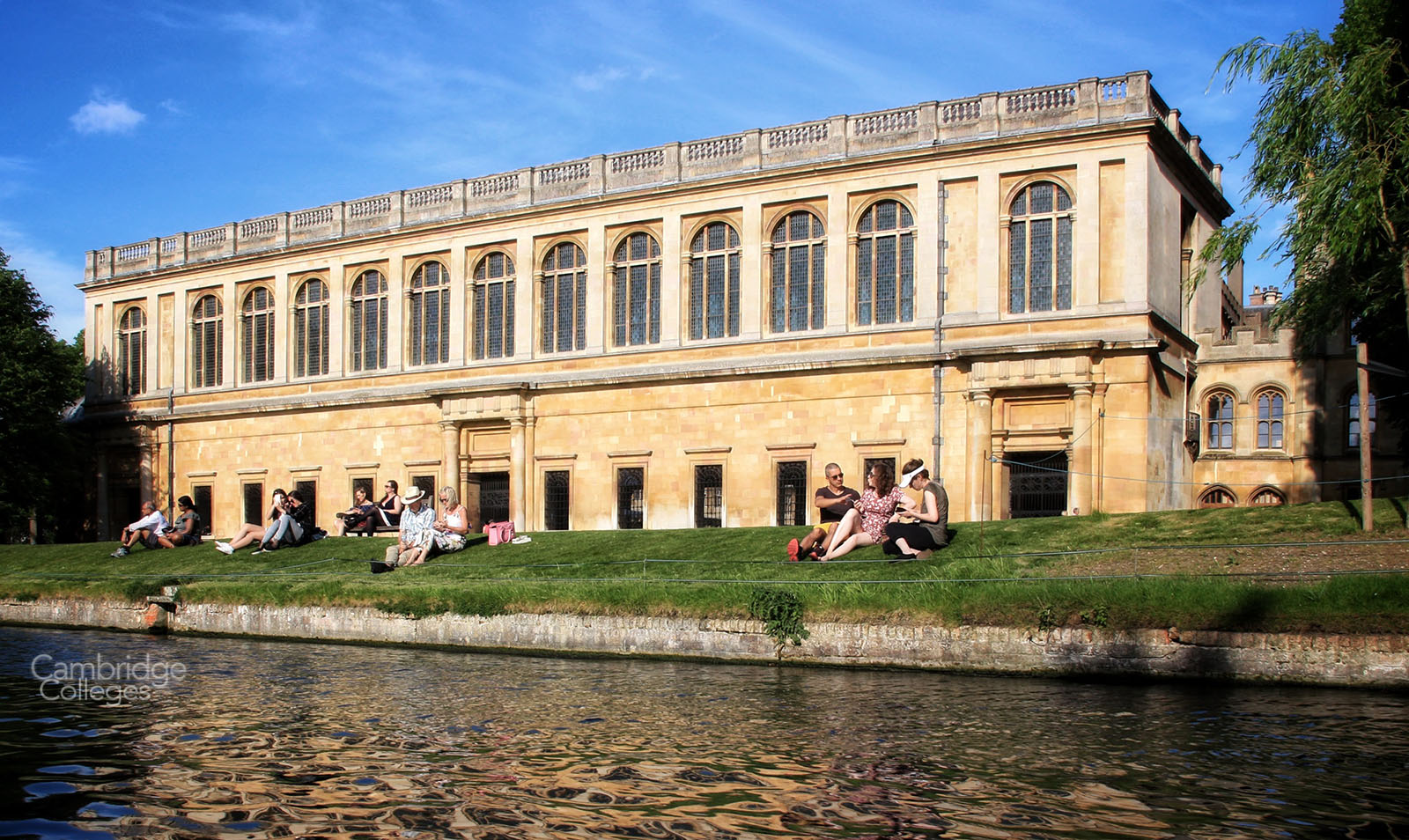 People sit on the bank of the river Cam outside the Wren Library on a summer day